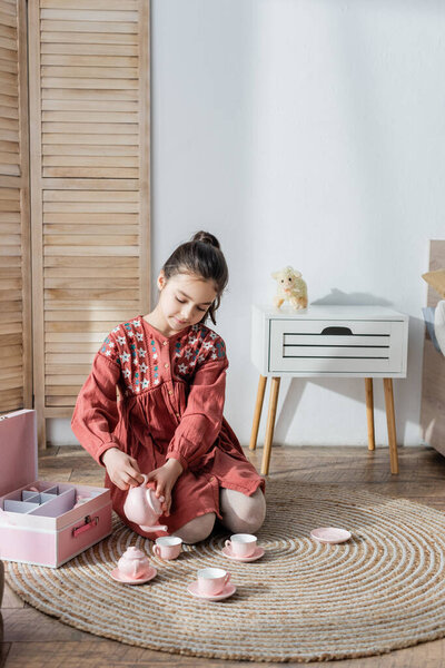 brunette girl sitting on floor carpet and pouring tea from toy teapot