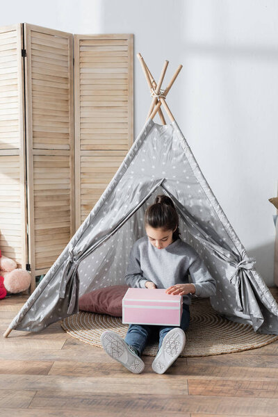 full length view of girl sitting on floor in wigwam and opening gift box