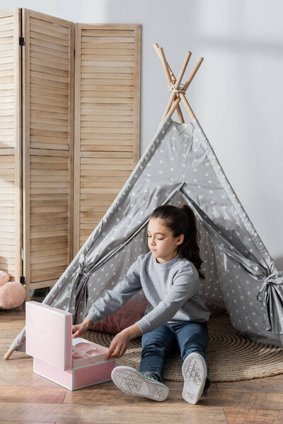 full length view of girl sitting in wigwam near box with toy tea set