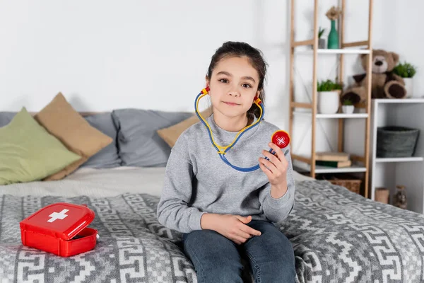Mädchen Mit Spielzeug Stethoskop Sitzt Auf Bett Neben Verbandskasten — Stockfoto