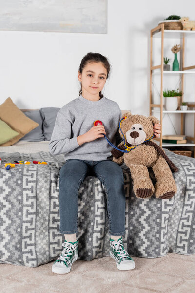 girl looking at camera while playing with teddy bear and toy stethoscope in bedroom