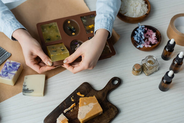 Top view of craftswoman taking soap from silicone mold near flowers and essential oils on table 