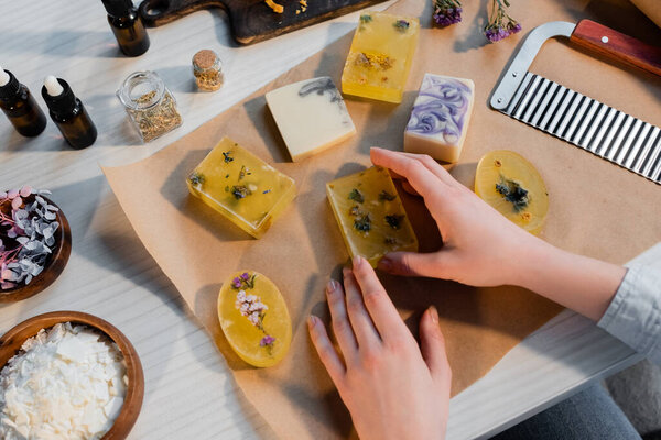 Top view of craftswoman holding handmade soap on craft paper near cutter and flowers on table 