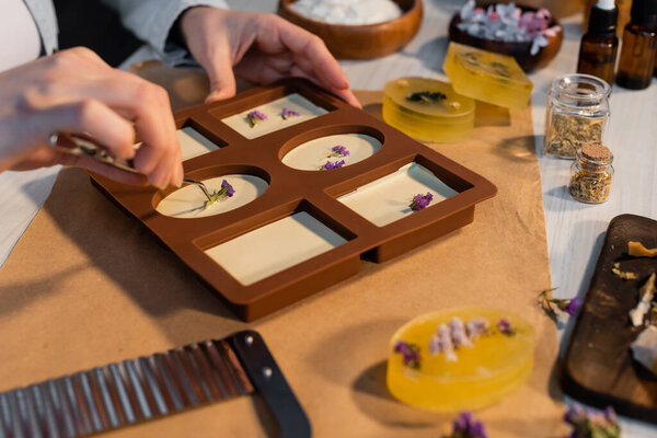 Cropped view of craftswoman putting dry flowers on craft soap in silicone mold at home 