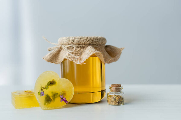 Jar with honey near handmade soap bars and herbs on table on grey background 