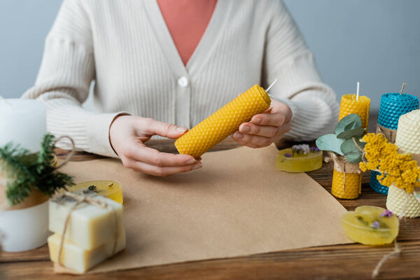 Cropped view of craftswoman holding handmade candle near soap and craft paper on table 
