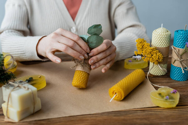 Cropped view of craftswoman decorating handmade candle with eucalyptus at home 