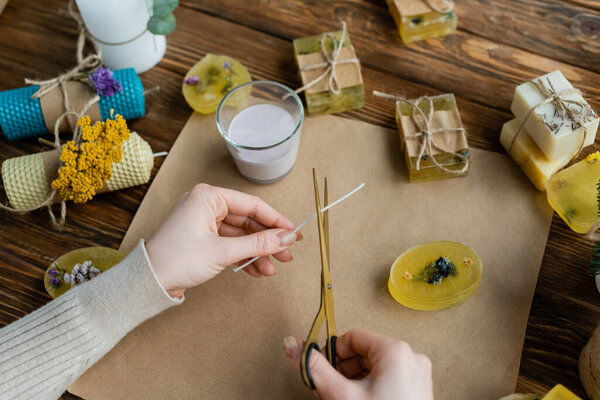 Top view of craftswoman cutting wick near handmade candles and soap at home 