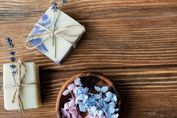 Top view of flowers in bowl near handmade soap bars on wooden surface 