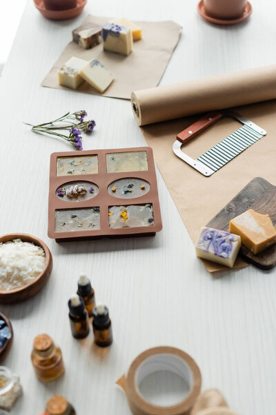 Soap bars in silicone mold near cutter, craft paper and essence oils on table 