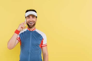 Spor giyimli ve vizörlü olumlu bir adam sarı alanda cep telefonuyla konuşuyor. 