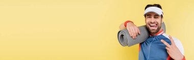 Young sportsman showing peace sign and holding fitness mat isolated on yellow, banner  clipart