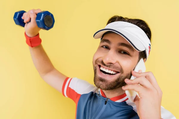 Smiling sportsman talking on smartphone and looking at camera while training with dumbbell isolated on yellow