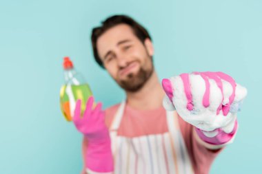 Plastik eldivenli bulanık bir adam. Elinde deterjan ve sünger var. Mavi üzerinde köpük var. 