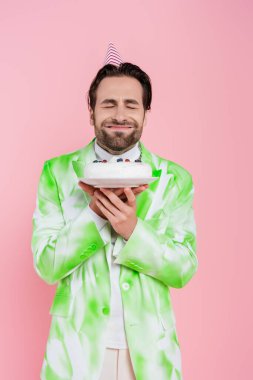 Smiling man in party cap closing eyes while holding birthday cake isolated on pink  clipart