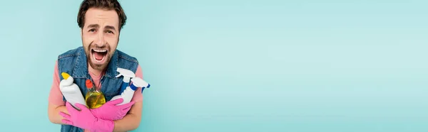 Młody Człowiek Gumowych Rękawiczkach Krzyczy Trzymając Detergenty Odizolowane Niebiesko Sztandar — Zdjęcie stockowe