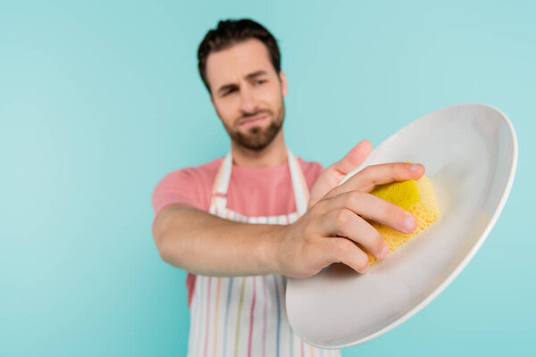 Blurred unmarried man in apron cleaning plate with sponge isolated on blue 
