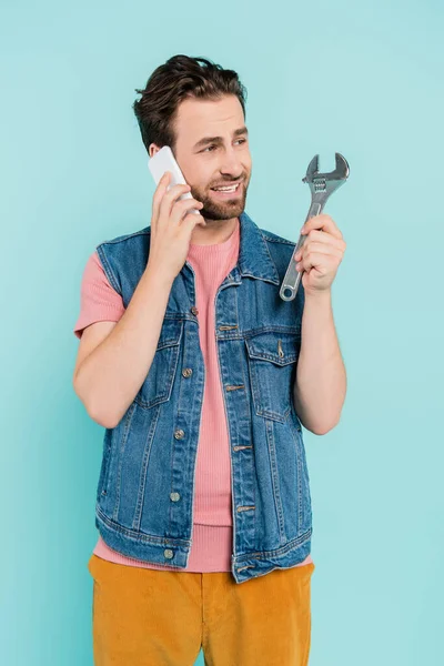 Positieve Man Praten Smartphone Kijken Naar Moersleutel Geïsoleerd Blauw — Stockfoto