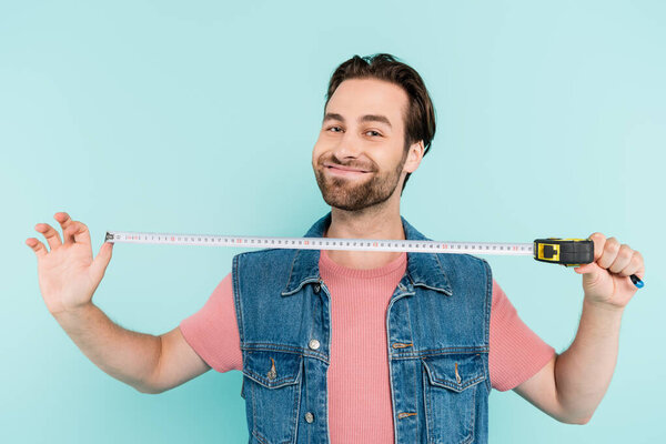 Cheerful man in denim vest holding tape measure isolated on blue 