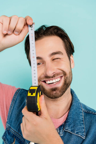 Smiling man in denim vest holding blurred tape measure isolated on blue 