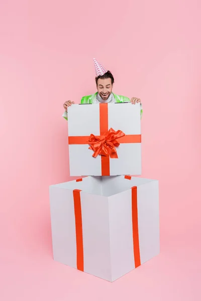 Excited man in party cap looking at huge gift box on pink background