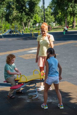 pleased woman standing near children and seesaw in park clipart