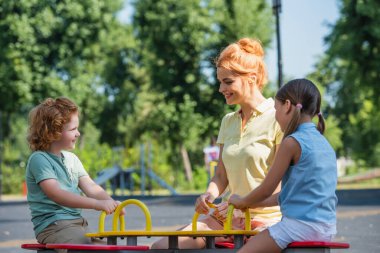 cheerful woman riding seesaw with children in summer park clipart