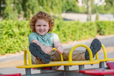 full length view of happy redhead boy riding seesaw in summer park clipart