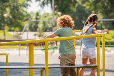 back view of brother and sister having fun on carousel in amusement park clipart