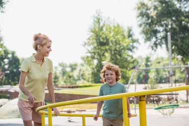 cheerful redhead woman smiling near son on carousel in park clipart