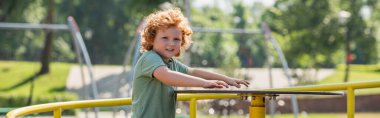redhead kid looking at camera while riding carousel in summer park, banner clipart