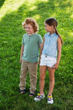 full length view of brother and sister in summer clothes standing on green lawn clipart