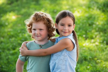 smiling girl embracing brother and looking at camera outdoors clipart