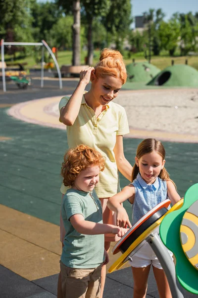 Mulher Sorridente Ajustando Cabelo Perto Crianças Divertindo Parque Infantil — Fotografia de Stock