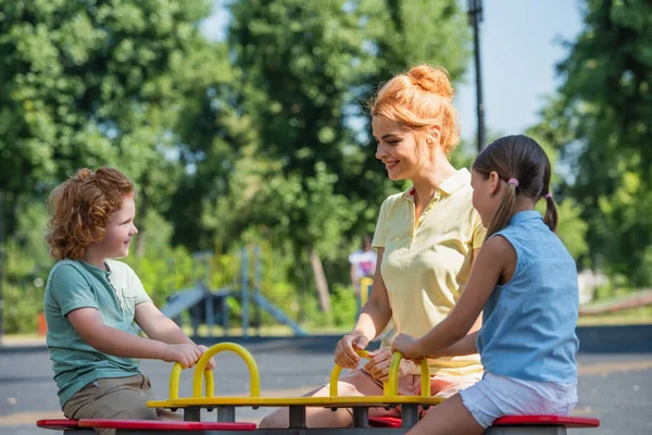 Neşeli kadın çocuklarla birlikte yaz parkında tahterevalliye biniyor.