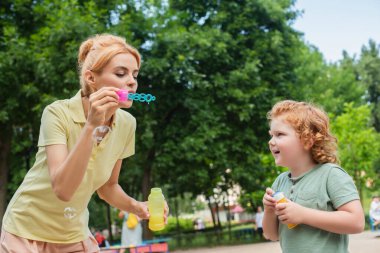 Kızıl saçlı kadın oyun parkında heyecanlı oğlunun yanında sabun köpüğü üflüyor.