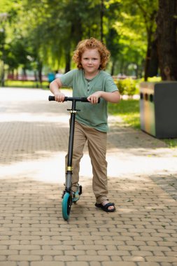 Yeşil parktaki kameraya bakan, scooter 'ı olan mutlu çocuk manzaralı.
