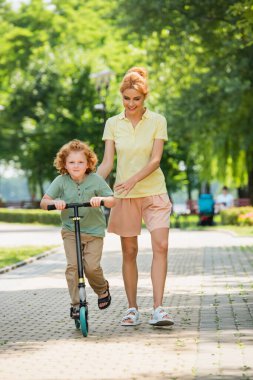 Parkta annemin yanında uçan scooter süren kızıl saçlı çocuğun uzun görüntüsü.