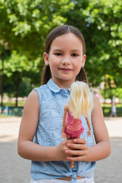 happy girl looking at camera while standing with doll in park