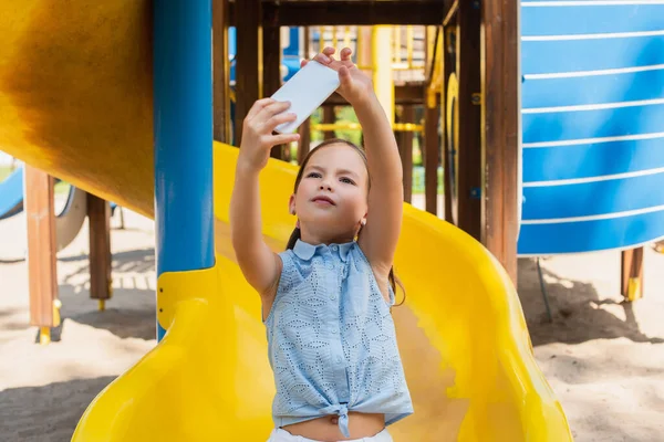 girl in sleeveless blouse taking selfie on slide in amusement park