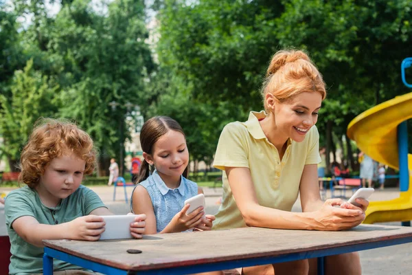 Χαρούμενη Οικογένεια Που Χρησιμοποιεί Smartphones Στην Παιδική Χαρά Στο Πάρκο — Φωτογραφία Αρχείου