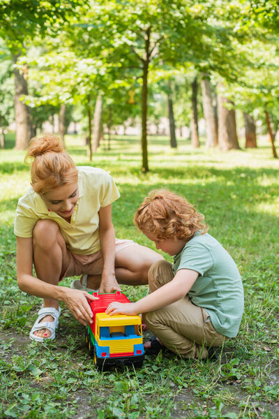 redhead mother and son playing with toy truck on green lawn in summer park