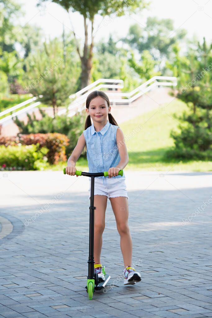 full length view of smiling girl riding on kick scooter in park and smiling at camera 