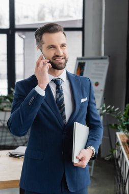 Neşeli ekonomist dizüstü bilgisayarını tutuyor ve bulanık ofisinde akıllı telefonla konuşuyor.