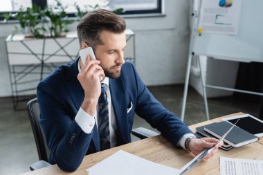 İş yerinde akıllı telefonla konuşurken belgelere bakan bir ekonomist.