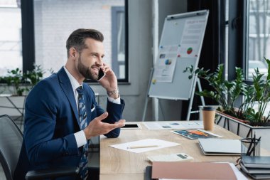 gülümseyen ekonomist akıllı telefonla konuşuyor. Ofiste kağıtların ve aletlerin yanında.