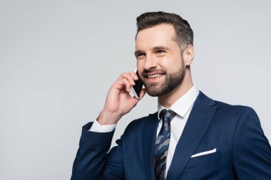 Mavi ceketli mutlu ekonomist gri renkte izole edilmiş akıllı telefonuyla konuşurken yüzünü çeviriyor.