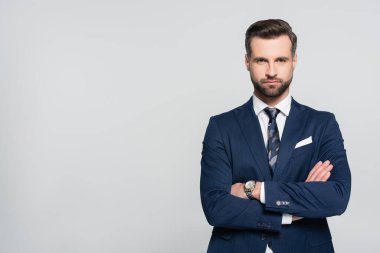 Mavi ceketli sakallı işadamı kollarını kavuşturup gri renkte duruyor.