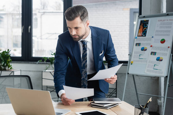 brunette economist holding papers and looking at laptop in office