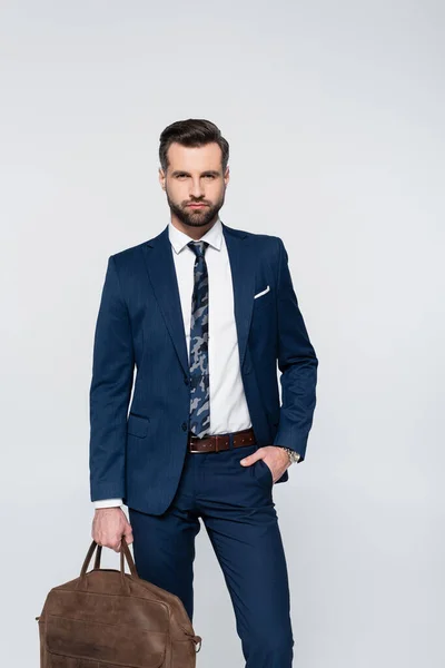 businessman in blue suit holding leather briefcase while standing with hand in pocket isolated on grey